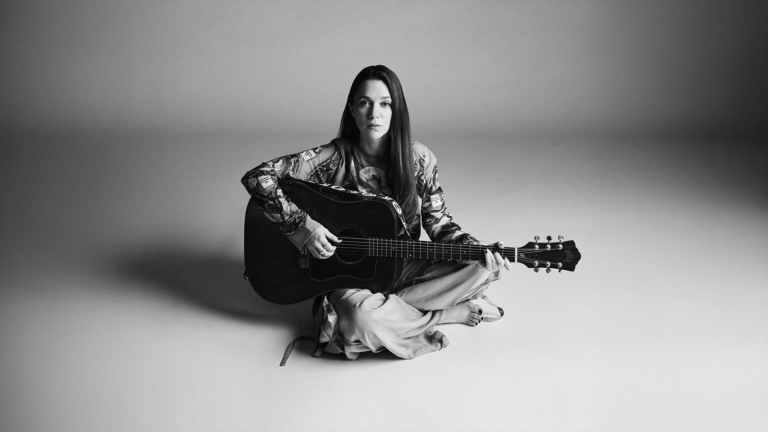 En kvinna sitter barfota på golvet i en ljus studio, iklädd mönstrad blus och vida byxor. Hon håller en akustisk gitarr i knät och spelar, samtidigt som hon tittar rakt in i kameran. Bilden är i svartvitt.